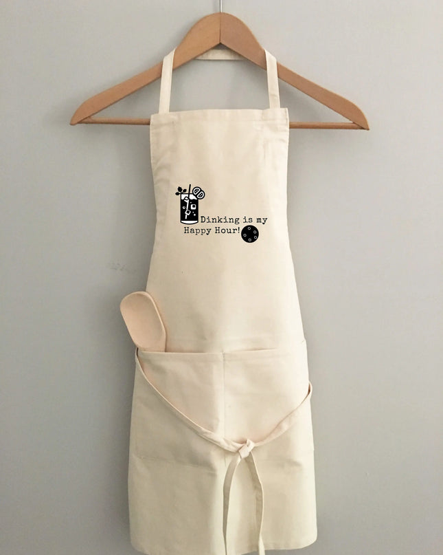 A white cotton apron with a pickleball and text graphic hanging on a wooden hanger.